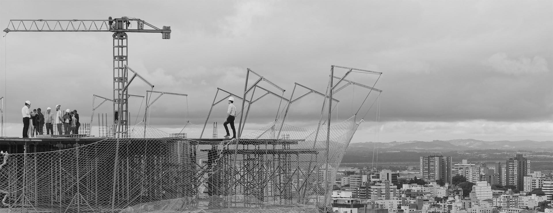 Gruas sobre uma obra da Colla, em preto e branco, com a vista de Porto Alegre ao fundo.