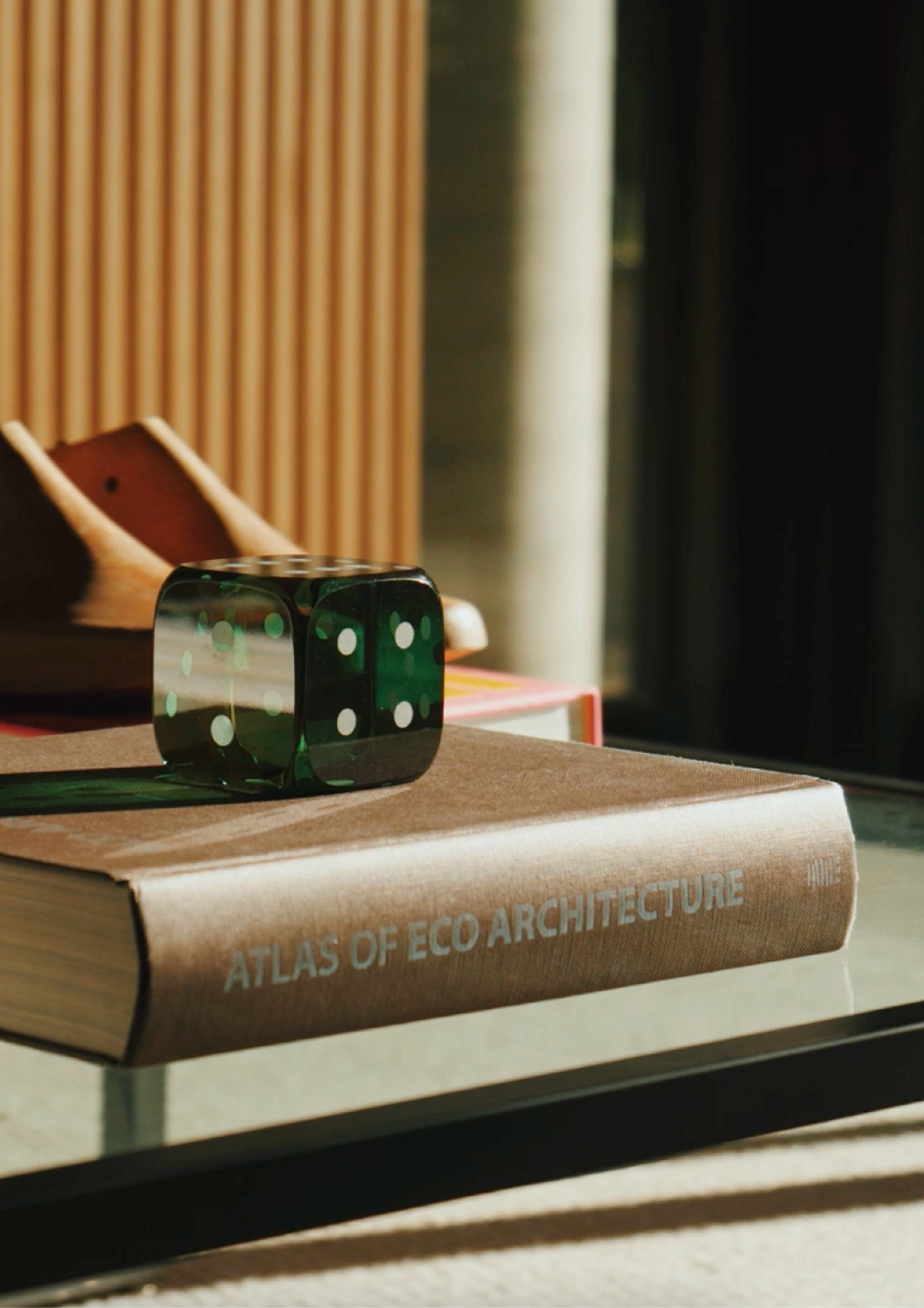 Livro Atlas of Eco Architecture sobre mesa, com peça de madeira e objeto verde ao lado.